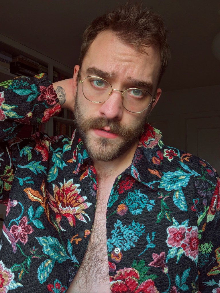 Hayden Ira May, a gay deaf man, wears a floral patterned shirt with pink and green flower patterns emblazoned on a dark colour. He stands holding one hand at the back of his neck. He is w wearing glasses and is looking at the camera.