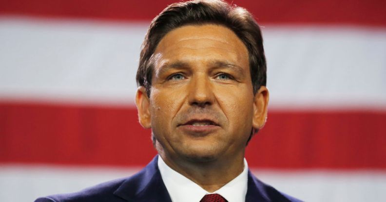 A photo of Florida governor Ron DeSantis wearing a dark suit, white shirt and red tie standing in front of the American flag