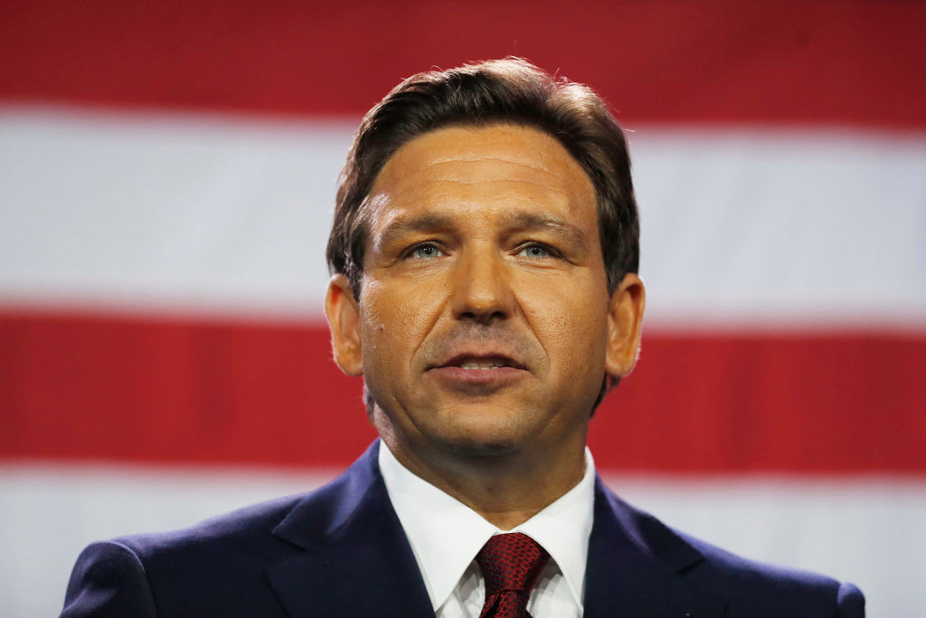 A photo of Florida governor Ron DeSantis wearing a dark suit, white shirt and red tie standing in front of the American flag
