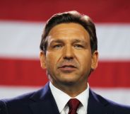 A photo of Florida governor Ron DeSantis wearing a dark suit, white shirt and red tie standing in front of the American flag