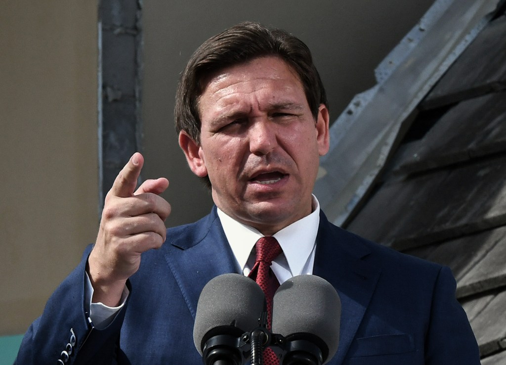 Florida governor Ron DeSantis wearing a navy suit, white shirt and red tie, stands in front of a microphone at a press conference