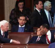 A photo shows US Republican George Santos wearing a dartk suit jacket over a white shirt and light blue sweater sitting alone in the House Chamber of the US Capitol building. All around him you can see other officials looking busy and talking to each other