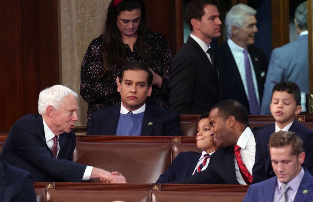 A photo shows US Republican George Santos wearing a dartk suit jacket over a white shirt and light blue sweater sitting alone in the House Chamber of the US Capitol building. All around him you can see other officials looking busy and talking to each other