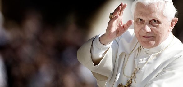 A photo of Pope Benedict XVI looking towards the camera and holding his hand up as if he's making a blessing