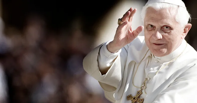 A photo of Pope Benedict XVI looking towards the camera and holding his hand up as if he's making a blessing