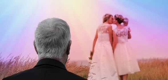 Two women kiss in wedding gowns while a man looks on