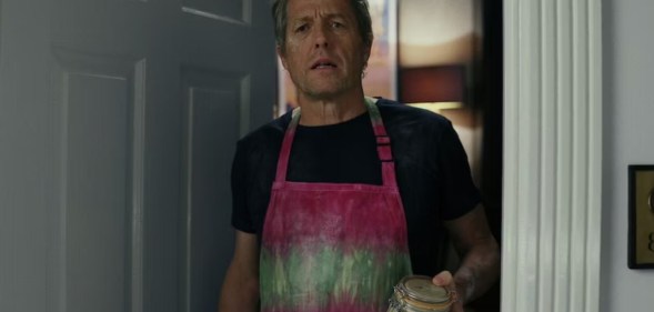 A still from the film Glass Onion: A Knives Out Mystery shows actor Hugh Grant wearing a pink and green apron over a black t-shirt as he stands in a doorway holding a glass jar of sourdough culture. (Netflix)