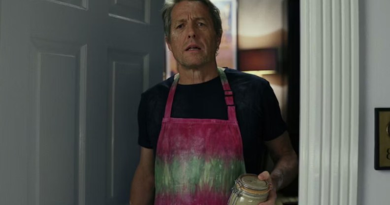 A still from the film Glass Onion: A Knives Out Mystery shows actor Hugh Grant wearing a pink and green apron over a black t-shirt as he stands in a doorway holding a glass jar of sourdough culture. (Netflix)