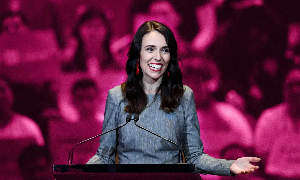 Jacinda Ardern is pictured standing at a podium speaking wearing a grey suit. The background is an edited pink one to show her LGBTQ+ support.