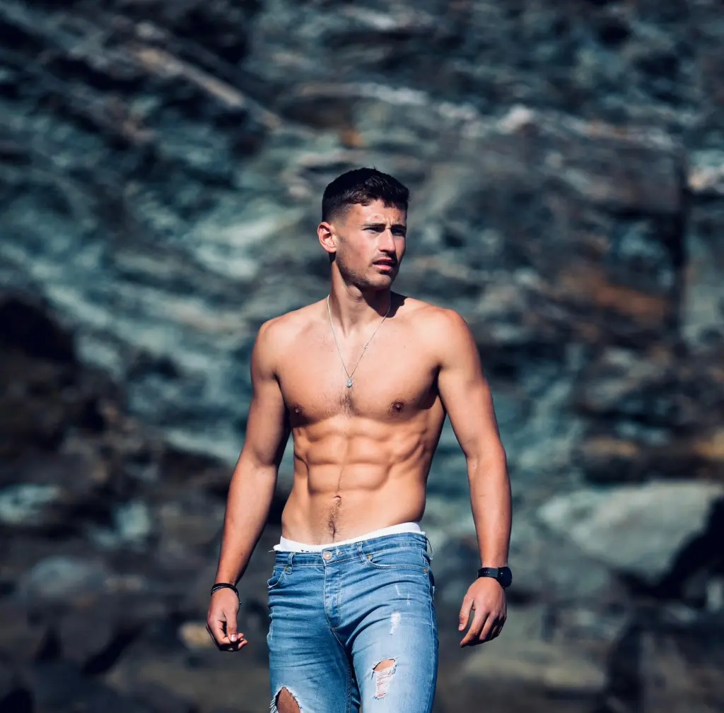 Jake Williamson pictured shirtless and wearing a pair of blue jeans by a cliff.