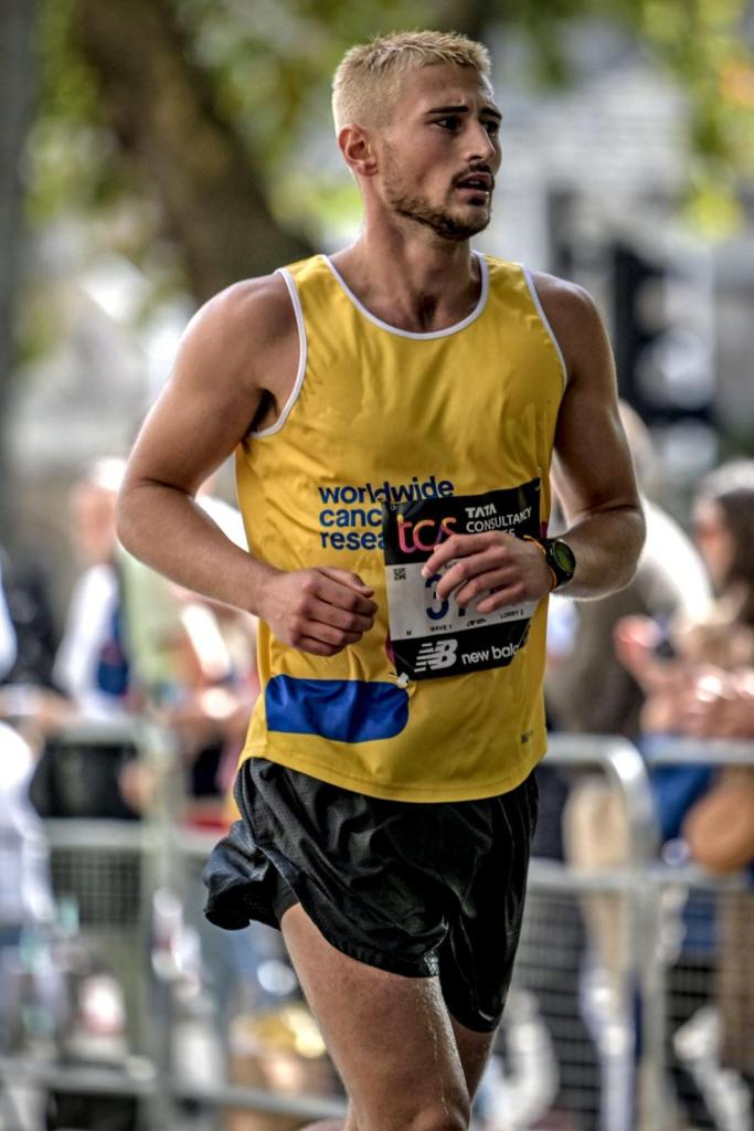 Jake Williamson running in a race. He is wearing a yellow vest and has blonde hair.