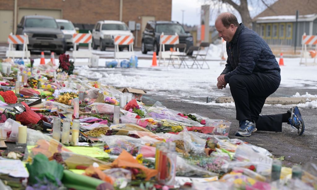 Jared Polis, wearing all black, kneels down in front of a huge array of flowers and sentimental gifts for the victims of the Club Q shooting.