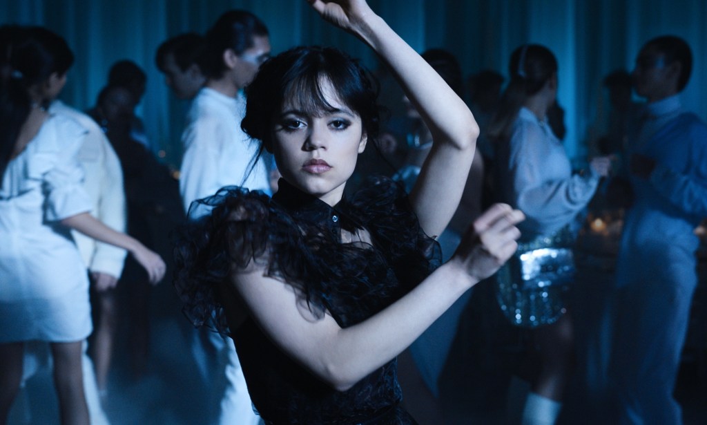 A screenshot from Netflix's Wednesday shows Jenna Ortega as Wednesday Addams dressed in black holding a pose as she dances - with other people also seen in the background dancing.