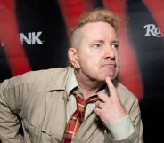 John Lydon wearing a beige jacket and checkered tie looking to the right with his finger on his chin.