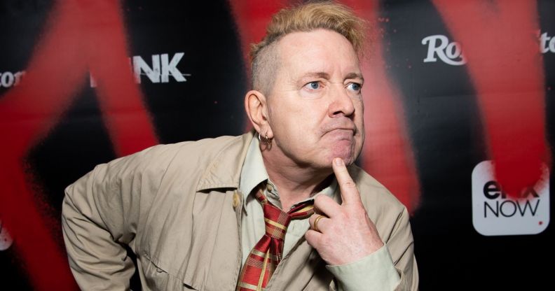 John Lydon wearing a beige jacket and checkered tie looking to the right with his finger on his chin.