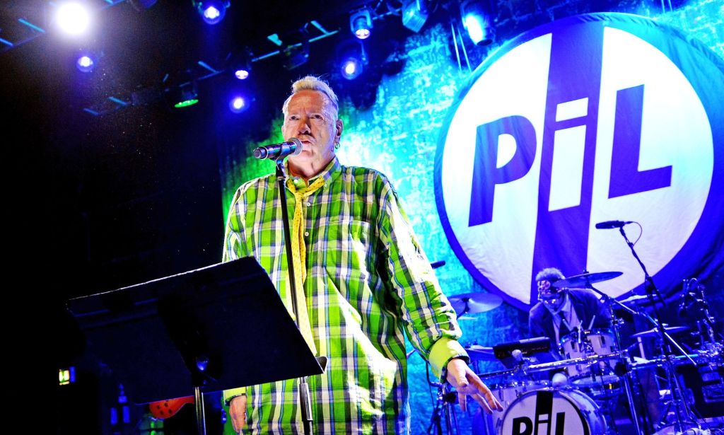 John Lydon wearing a green and yellow suit performing on stage with a microphone and music sheet stand, against a backdrop of his band's logo.