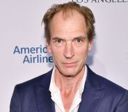 Julian Sands, infront of a white background, smiles on the red carpet.