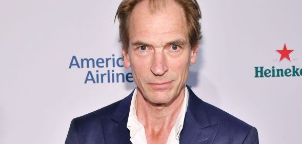 Julian Sands, infront of a white background, smiles on the red carpet.