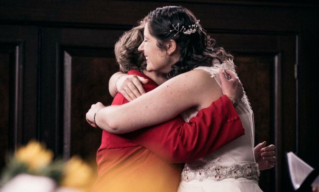 Sarah Barley-McMullen pictured marrying her wife Helen. Sarah and Helen are pictured embracing in a church. Sarah is wearing a red suit and Helen, on the right, is wearing a sleeveless white wedding gown.