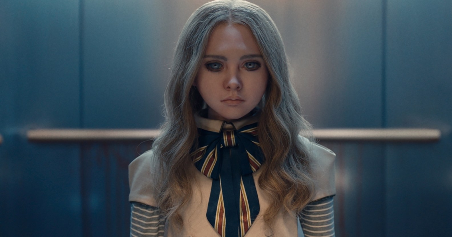 A still from the movie M3GAN showing the robot doll wearing a white and blue-striped outfit standing in an elevator