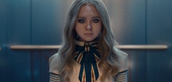 A still from the movie M3GAN showing the robot doll wearing a white and blue-striped outfit standing in an elevator