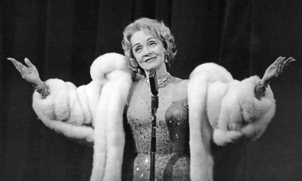 In this black and white photo, Marlene Dietrich wears a sparkly dress and a white fur coat as she performs on stage with both her arms held out to her sides