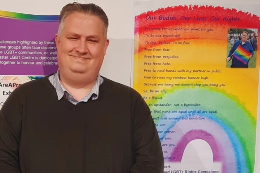 Mathew Hulbert pictured at an event. He is wearing a dark coloured jumper and shirt and a graphic showing the rainbow colours is visible in the background.