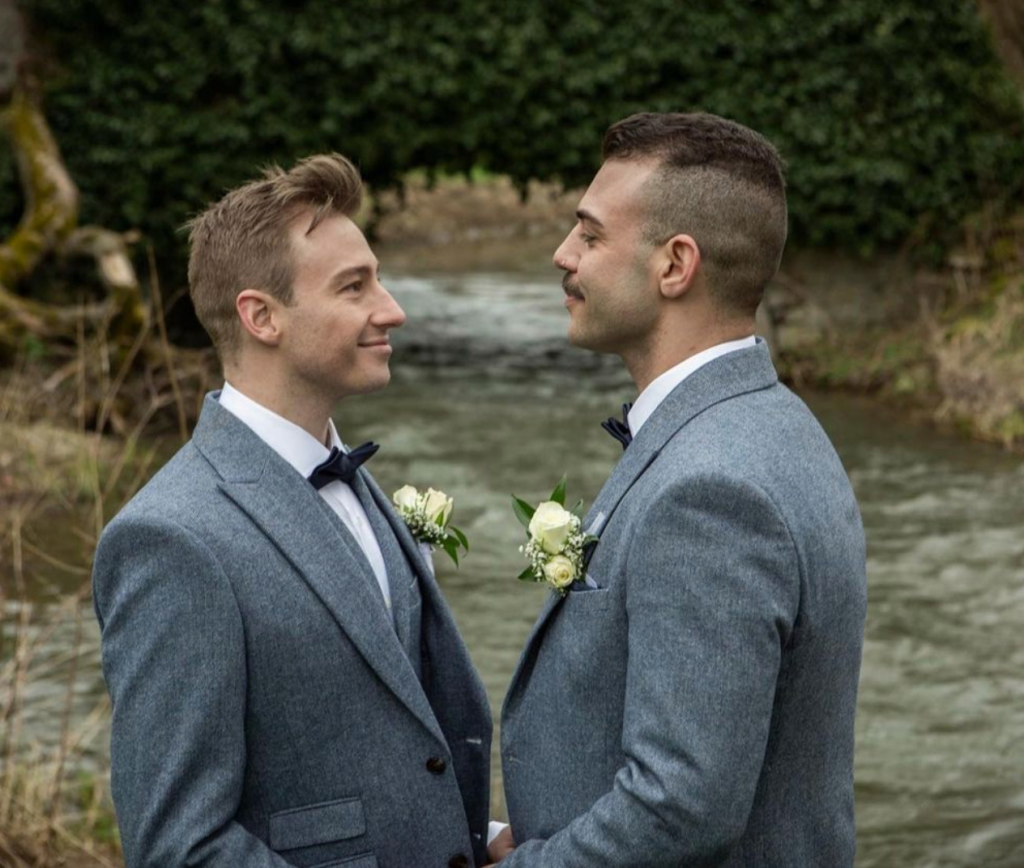 Matthew Mitcham pictured outdoors with his husband Luke Rutherford on their wedding day. Matthew is pictured on the left wearing a grey suit. Luke is pictured on the right wearing a similar garment.