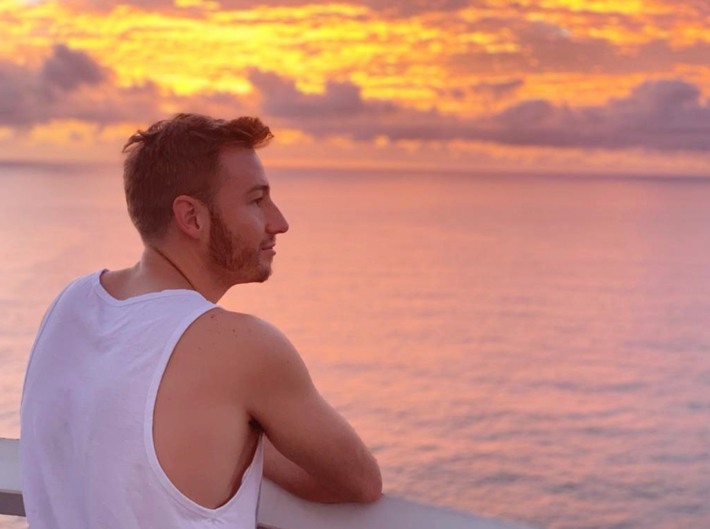 Matthew Mitcham staring out at the sun setting over the sea. He stands in the left foreground wearing a white vest.