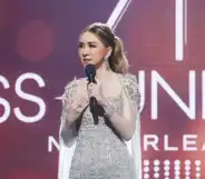 A still from the 71st Miss Universe pageant shows the franchise's owner, a trans woman called Jakkaphong "Anne" Jakrajutatip wearing a silver dress and holding a microphone standing on a stage with the "Miss Universe New Orleans" logo showing in the background
