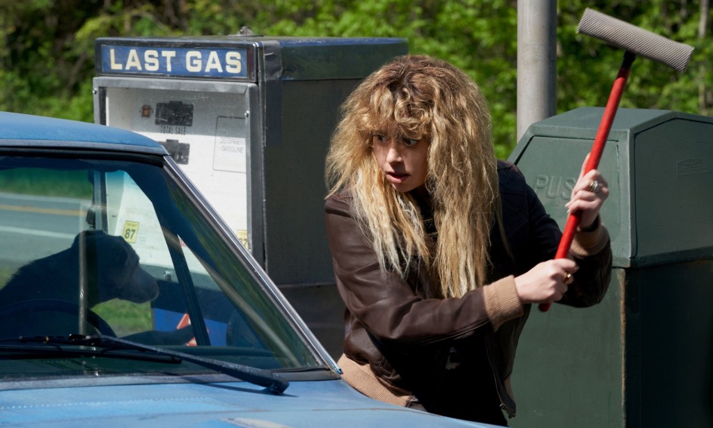 A screenshot of actor Natasha Lyonne as Charlie Cale in Poker Face In the scene she is seen wielding a squidgy mop at a car that has a dog sitting at the window.