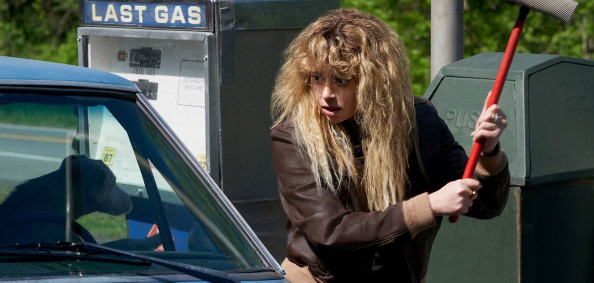A screenshot of actor Natasha Lyonne as Charlie Cale in Poker Face In the scene she is seen wielding a squidgy mop at a car that has a dog sitting at the window.