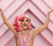 Drag queen Panti Bliss wears a pink and patterned dress with her hands raised in the air. She has pink hair on the left side and blonde on the right. She is standing against a pink patterned background in a promotional shot for the RTÉ show Dancing with the Stars.