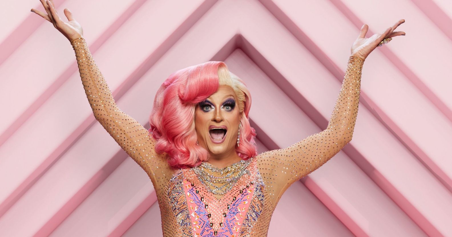 Drag queen Panti Bliss wears a pink and patterned dress with her hands raised in the air. She has pink hair on the left side and blonde on the right. She is standing against a pink patterned background in a promotional shot for the RTÉ show Dancing with the Stars.