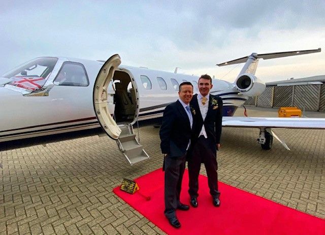 Paul and Alain on their wedding day. They are both wearing suits and are standing on a red carpet outside a private jet.