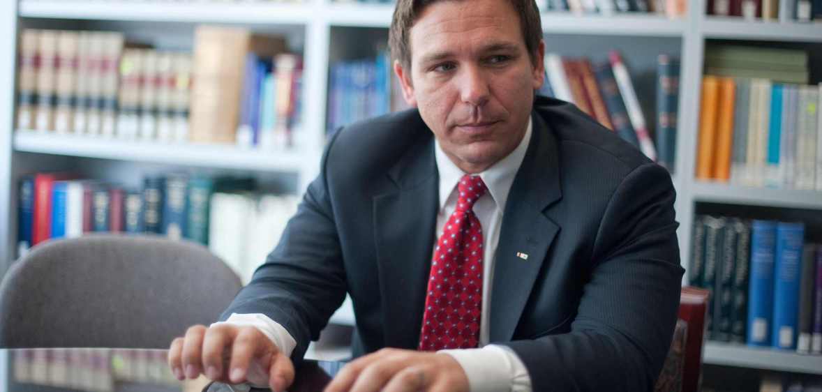A photo of Florida governor Ron DeSantis wearing a dark suit, white shirt and red tie standing in a room with the background showing books on shelves