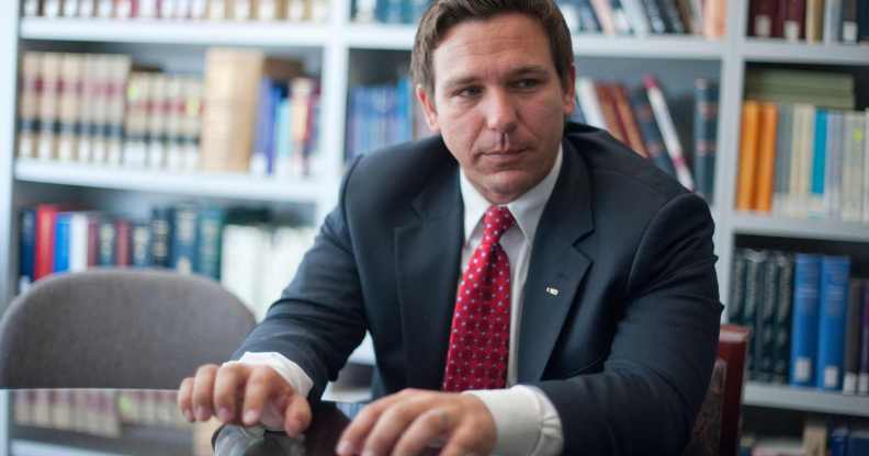 A photo of Florida governor Ron DeSantis wearing a dark suit, white shirt and red tie standing in a room with the background showing books on shelves