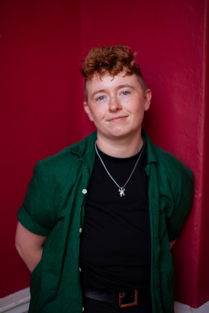 Ruadhán Ó Críodáin, executive director of ShoutOut, an Irish LGBTQ+ charity. He is pictured wearing a green shirt and a black t-shirt with a gold chain around his neck. he is standing against a dark red background.