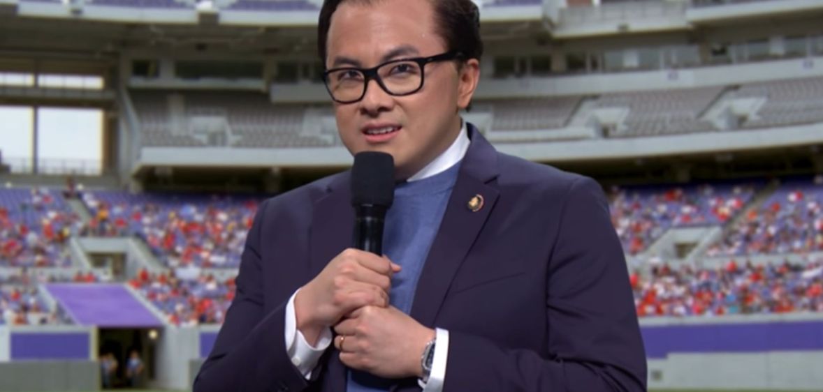 Bowen Yang dressed as George Santos during an SNL skit infront of a greenscreen of an NFL football field.