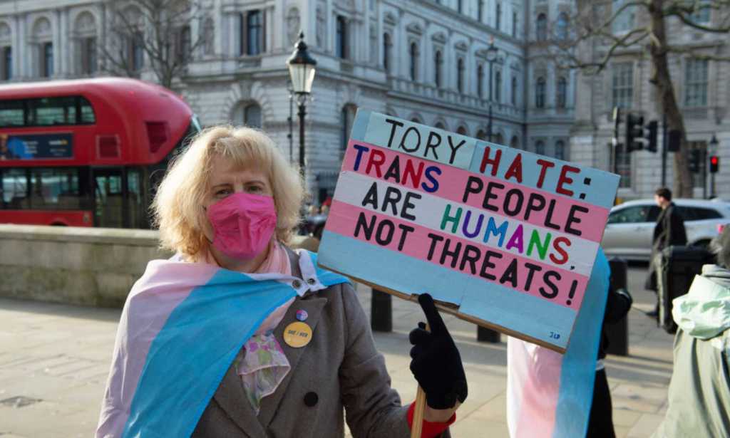 A trans activist takes part in a protest opposite Downing Street on 17 January 2023. The person is holding a sign that reads: "Tory hate: Trans people are humans not threats!" the sign is coloured in the colours of the trans flag, and the person is wearing a trans Pride flag draped around them.