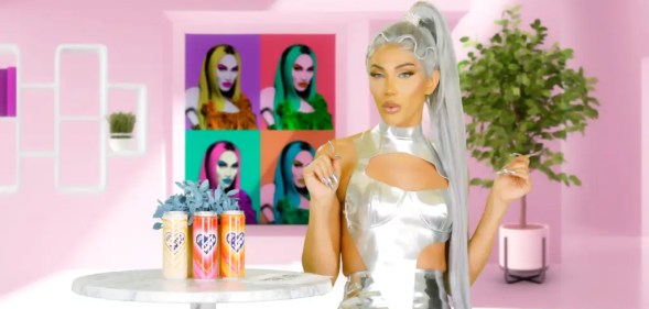 A promo image for new series Kerri Kares shows drag queen Kerri Colby dressed in a white frilly dress and rainbow-coloured wig looking towards the camera while she is set against a pink and starry background