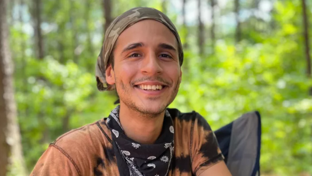 A photo of queer environmental activist Manuel “Tortuguita” Terán wearing a light orange t-shirt and smiling, with a forest in the background