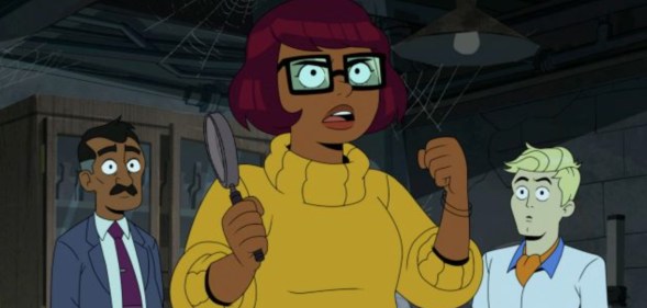 A still from new HBO Max cartoon Velma shows main character Velma Dinkley wearing a yellow sweater and holding magnifying glass in one hand while Fred dressed in white shirt and orange cravat is standing to the right. (HBO Max)