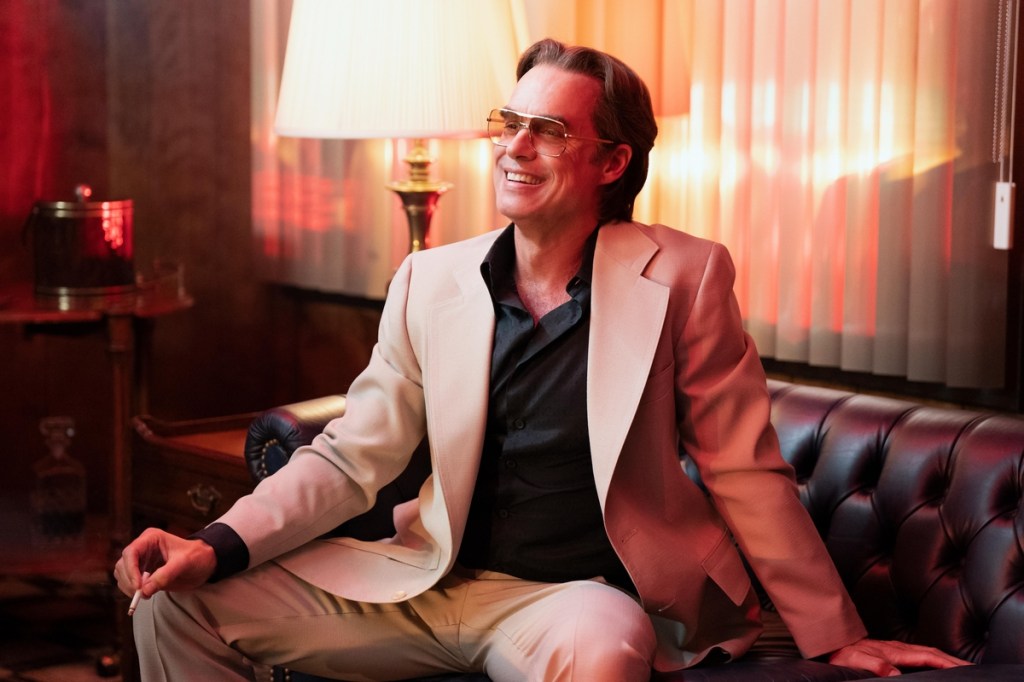A still from Disney+ series Welcome to Chippendales shows actor Murray Bartlett as character Nick de Noia dressed in a black shirt and cream suit wearing tinted glasses and sitting on a leather chair