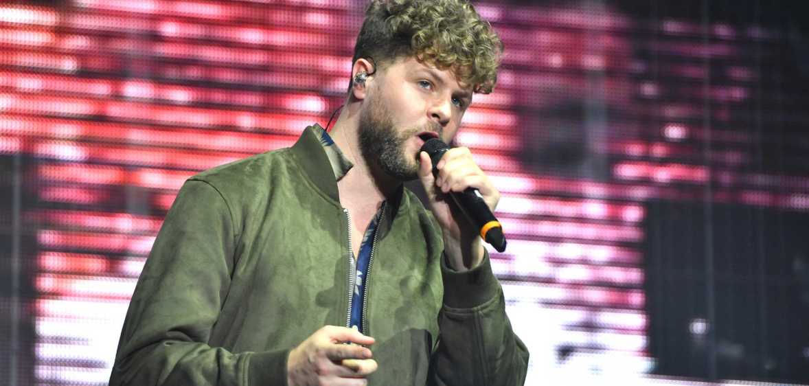 A photo of The Wanted's Jay McGuinness wearing a green top and singing into a microphone while performing on stage
