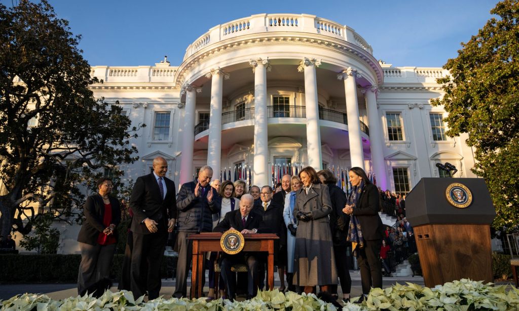 President Joe Biden signs the Respect for Marriage Act while surrounded by a group of people on the White House lawn with the White House lit up in rainbow LGBTQ+ colours