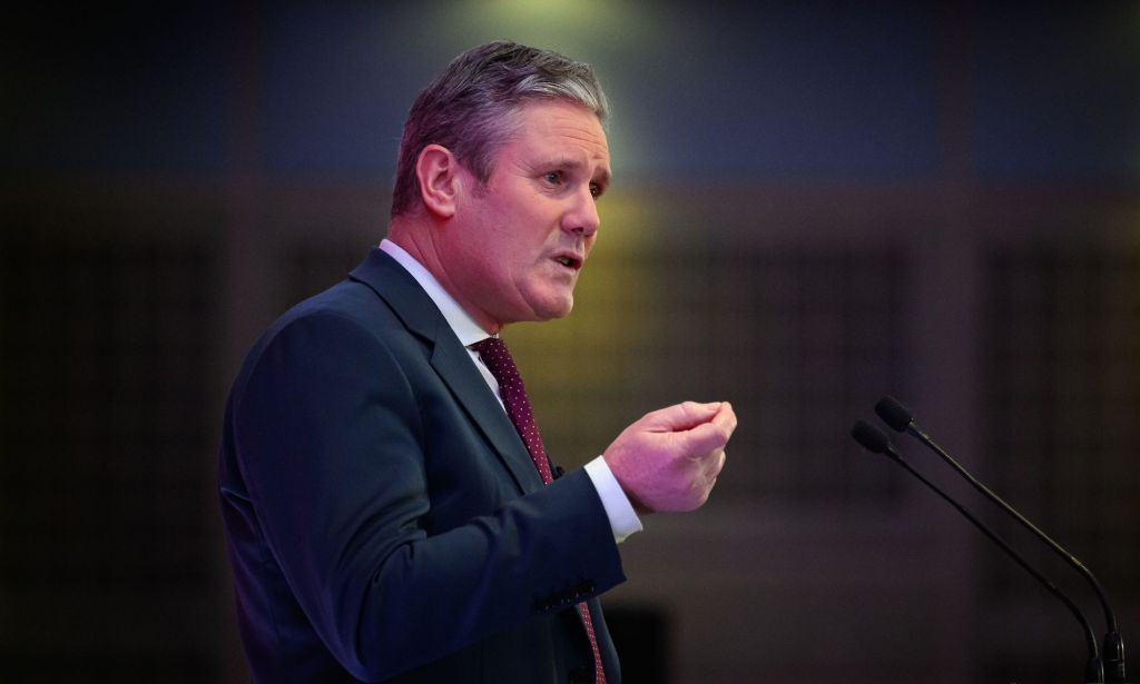 UK Labour Party leader Keir Starmer wears a white button-up shirt, red tie and blue jacket as he speaks at an event. He is standing at a podium and gesturing with one hand out