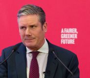 UK Labour Party leader Keir Starmer wears a white button-up shirt, red tie and blue jacket as he speaks at an event. He is standing at a podium in front of a red background