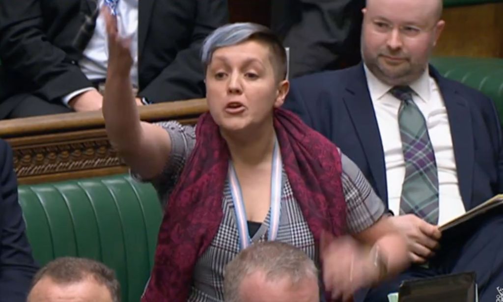 SNP MP Kirsty Blackman gestures with one hand while she wears a grey patterned outfit with a red scarf during a parliamentary discussion on why the UK government is blocking Scotland's Gender Recognition Reform bill from becoming law