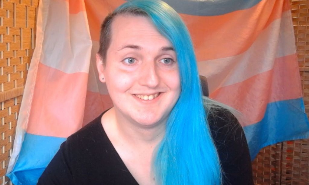 Laura Kate Dale wears a dark top. She has long, blue hair with an undercut. She is sitting in front of a trans Pride flag